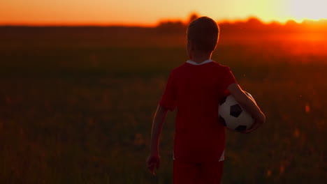 Young-football-player-goes-with-the-ball-on-the-field-dreaming-of-a-football-career-at-sunset-looking-at-the-sun.-The-camera-watches-the-boy-walking-on-the-field-at-sunset-with-a-soccer-ball.